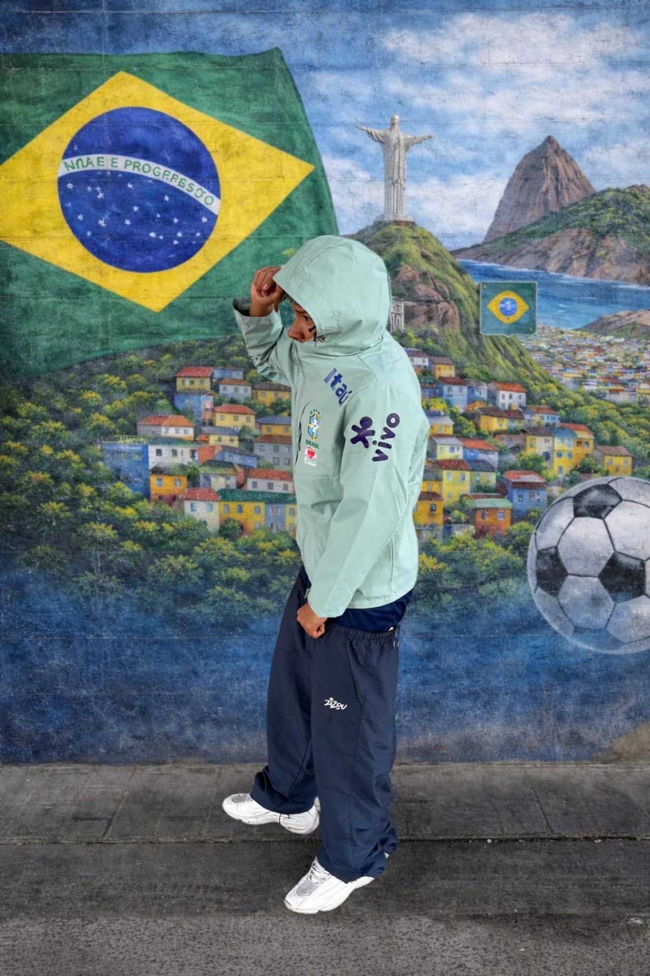 Veste du Brésil verte portée capuche relevée devant une fresque du drapeau brésilien et du Christ